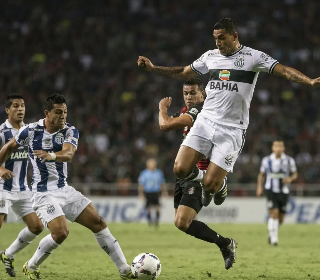Bahia precisa vencer o Santos para voltar ao G-8; transmissão ao vivo Jogador do Bahia chutando a bola de costas, zagueiros do Santos perseguindo, arquibancada ao fundo